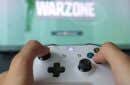 A person holding a white Xbox controller with the game title "WARZONE" displayed on a blurred TV screen in the background - person-holding-a-white-xbox-controller-warzone-game-title-on-a-blurred-tv-screen-in-the-background