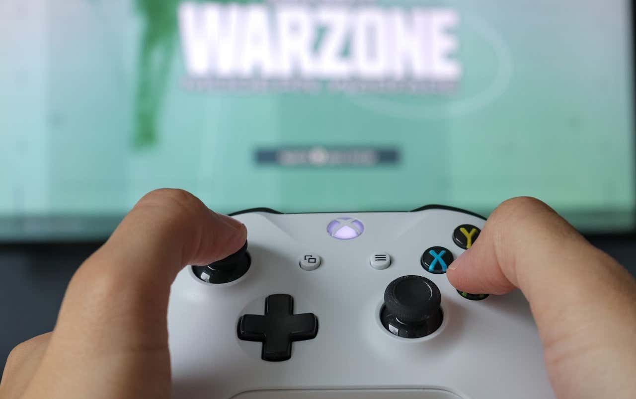 A person holding a white Xbox controller with the game title "WARZONE" displayed on a blurred TV screen in the background - person-holding-a-white-xbox-controller-warzone-game-title-on-a-blurred-tv-screen-in-the-background